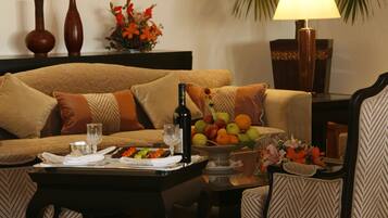 A living room with a sofa, coffee table, and two chairs. There is a lamp, a vase with flowers, and a fruit bowl on the table.