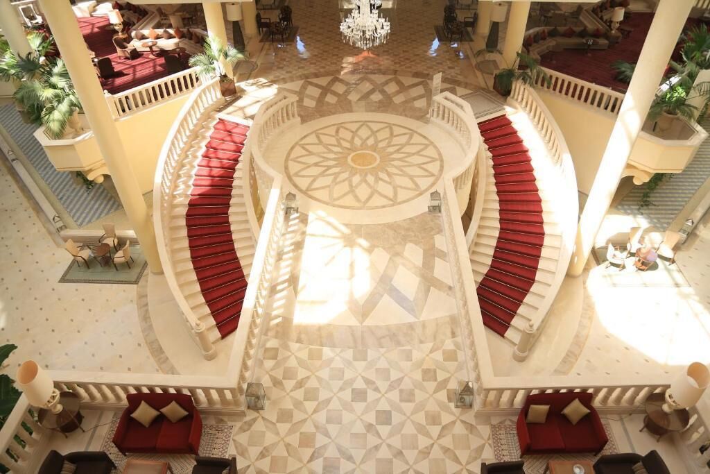 A grand lobby with a central staircase, red cushioned seating, and a patterned floor.