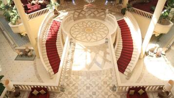 A grand lobby with a central staircase, red cushioned seating, and a patterned floor.
