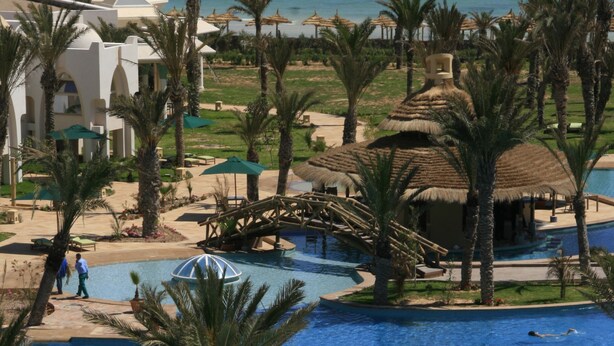 Ein Poolbereich eines Resorts mit Palmen, einer Brücke und Blick auf den Ozean.