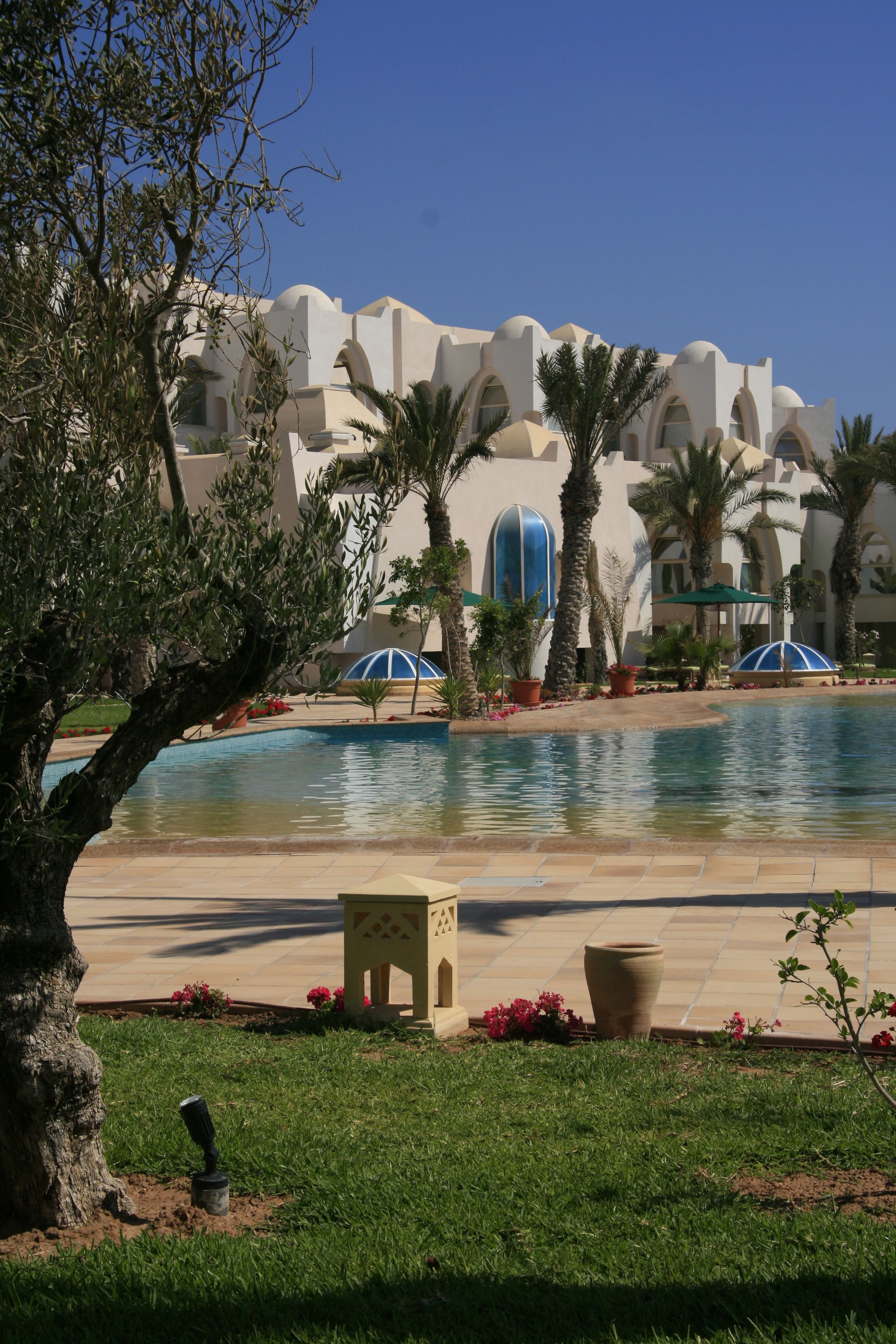 Un área de piscina con palmeras, un edificio grande con ventanas arqueadas y un cielo azul despejado.