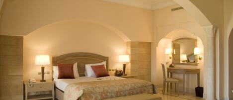 A hotel room with an arched ceiling, a bed with a headboard, bedside lamps, a bench, a desk with a chair, and a mirror.