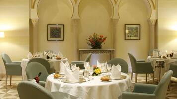 A dining area with a round table set for four, surrounded by four chairs. The table is covered with a white tablecloth, and there are various dishes and glasses on it. The room has a classic architectural design with arched doorways and a decorative ceiling.
