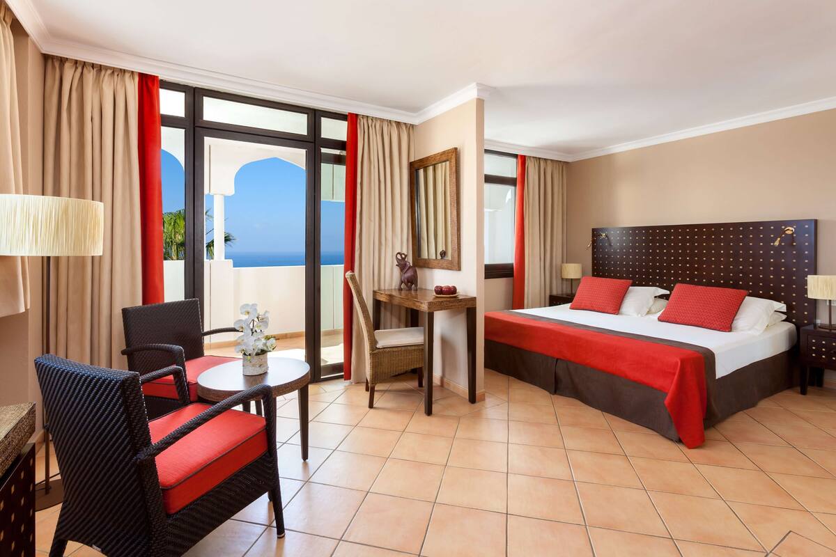 Quarto de hotel com cama, escrivaninha, cadeira e vista para o oceano.