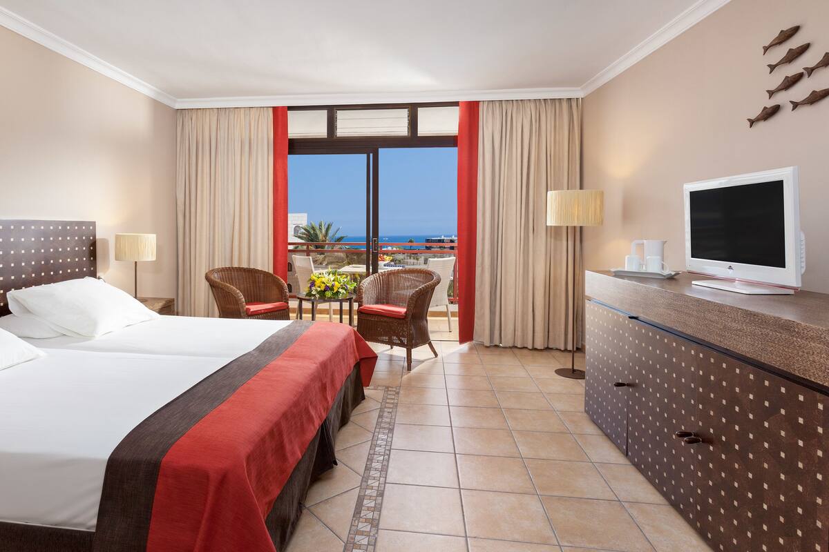 Quarto de hotel com cama, escrivaninha com televisão, cadeira e vista para o oceano.