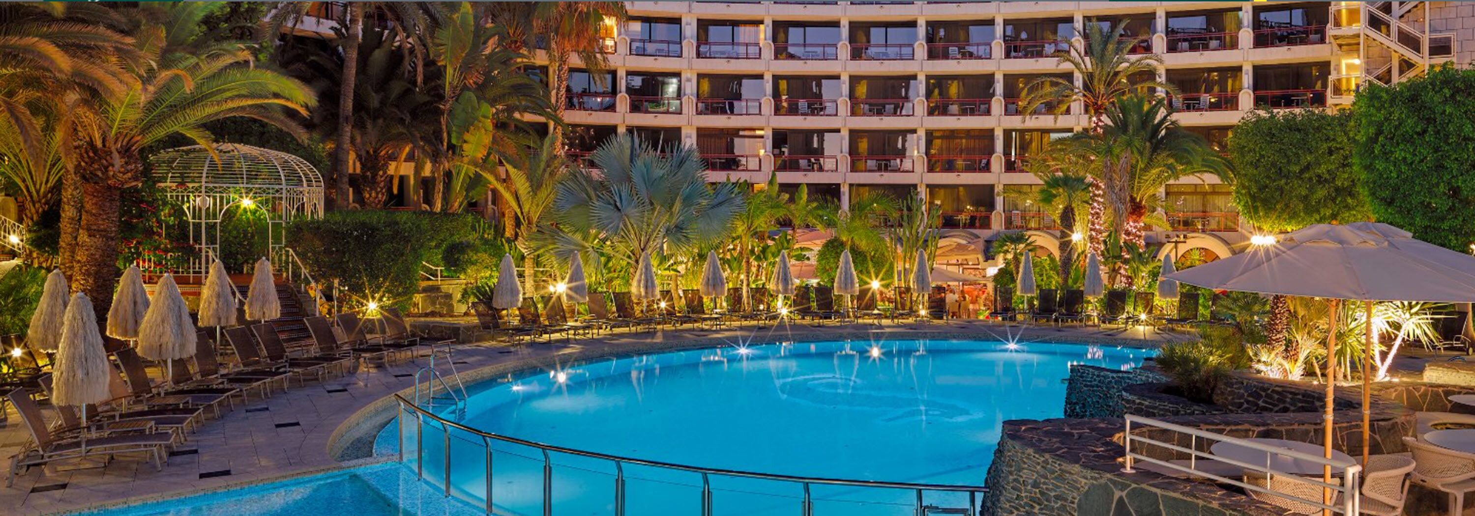 Zona de piscina de hotel con una piscina grande, sillas de descanso, sombrillas y un edificio de varios pisos al fondo.