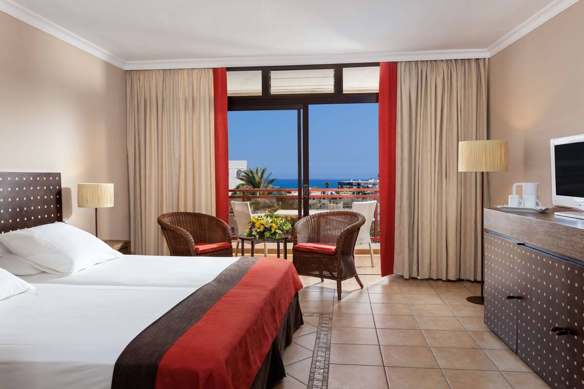 Quarto de hotel com duas camas, uma mesa de jantar com cadeiras, uma televisão e vista para o mar.