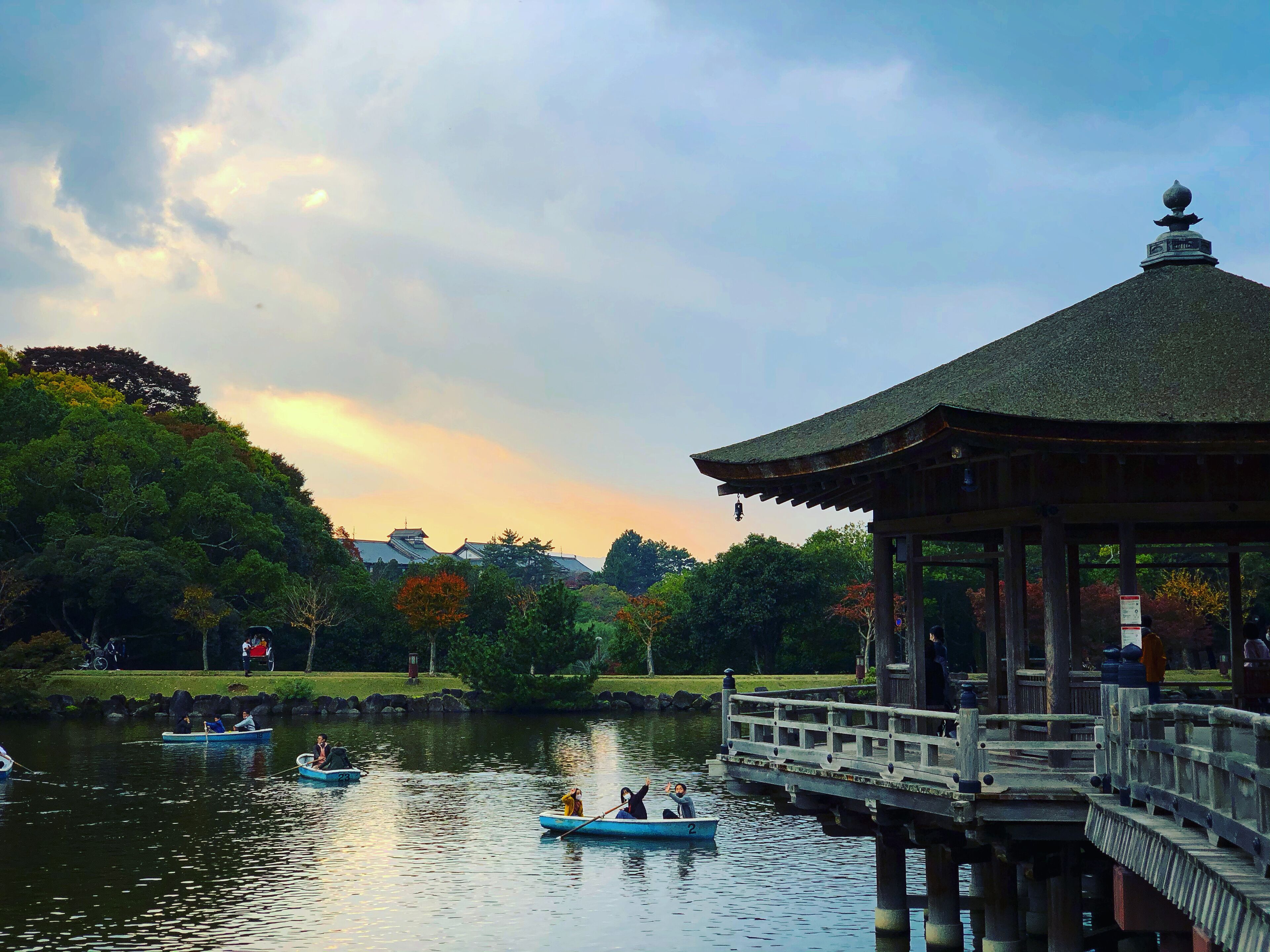 A traditional pavilion with a tiled roof, a wooden deck, and a view of a calm lake with boats.