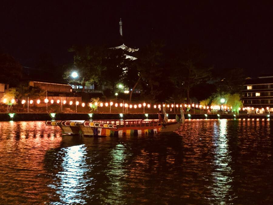 Escena nocturna de un río con un bote, árboles iluminados y una torre al fondo.