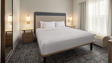 A hotel room with a large bed, two bedside tables, a nightstand, a window with sheer curtains, and a wall-mounted lamp.