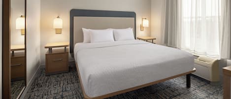 A hotel room with a large bed, two bedside tables, a nightstand, a window with sheer curtains, and a wall-mounted lamp.