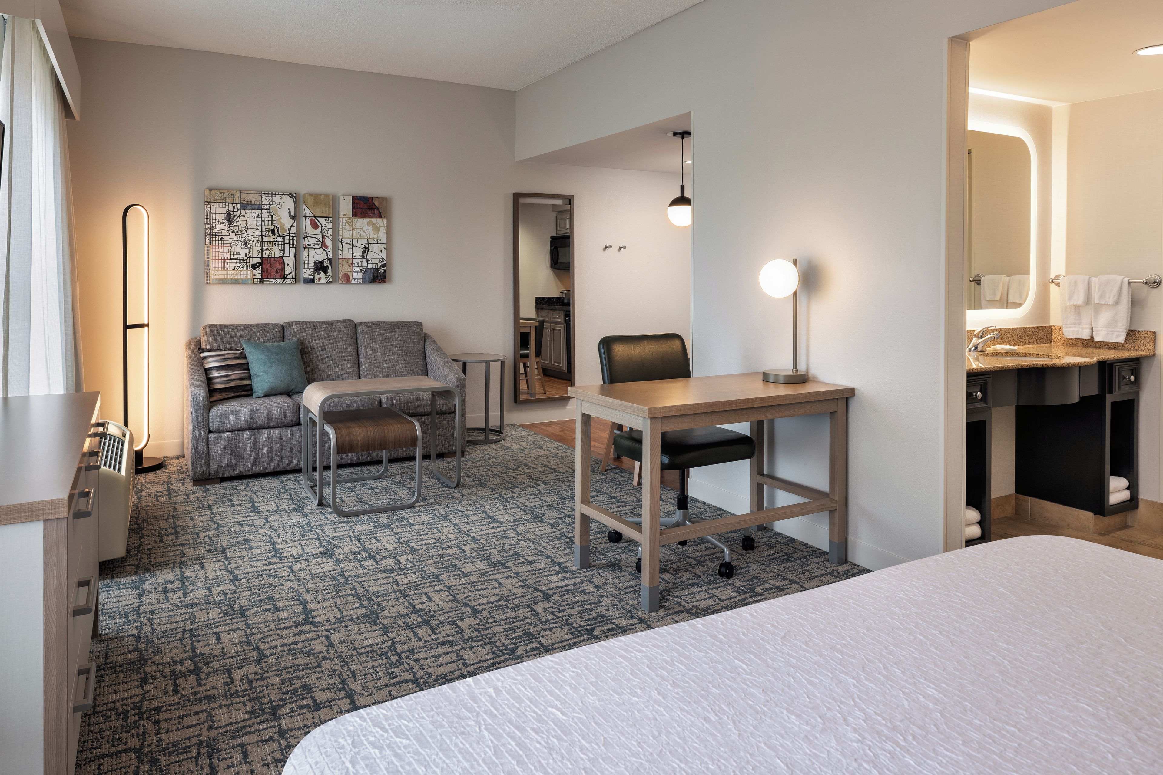 A hotel room with a bed, a desk, a sofa, a chair, a small table, a rug, a wall with artwork, a mirror, and a bathroom with a sink and towel rack.