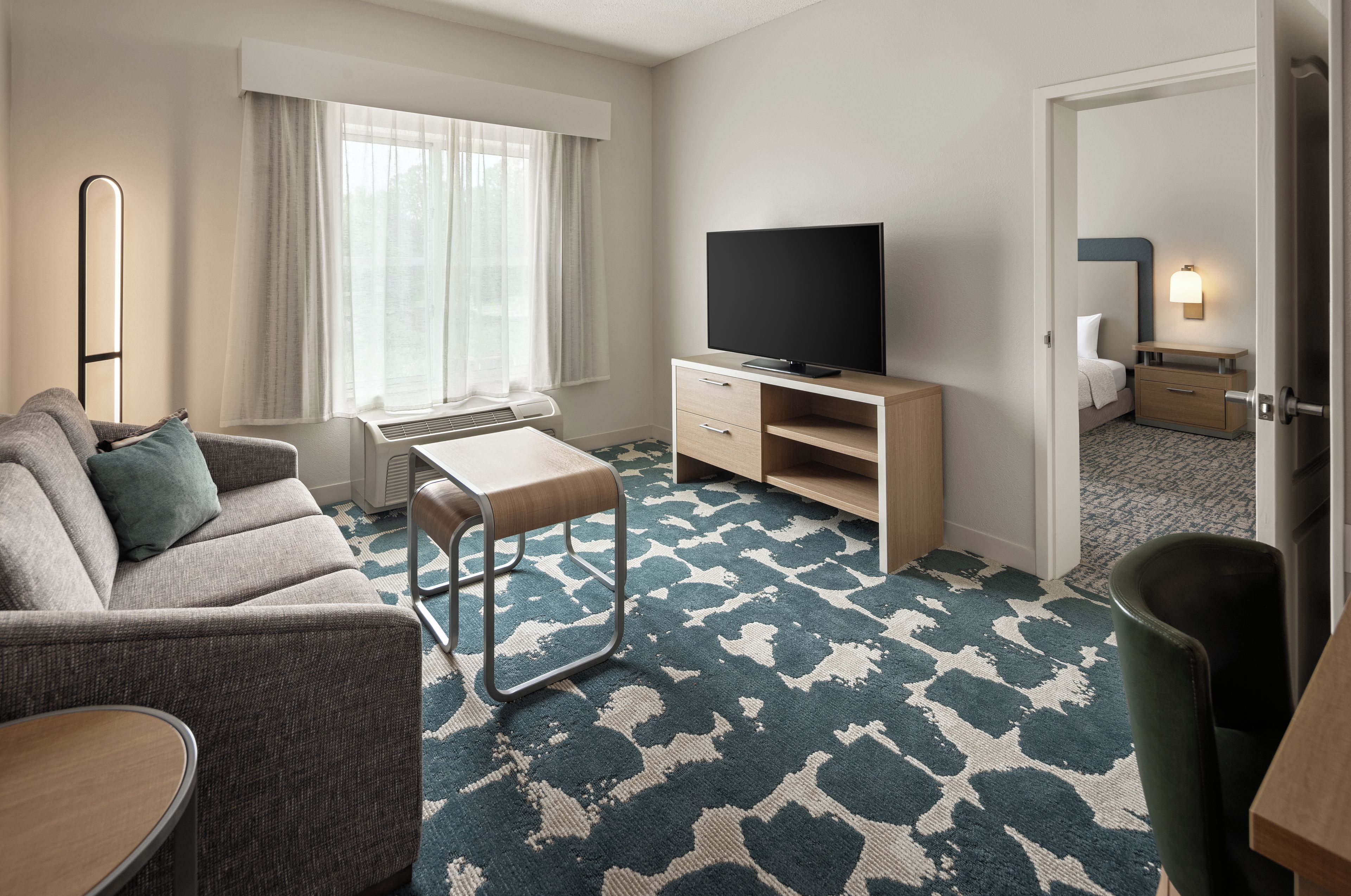 A hotel room with a sofa, a small table, a TV on a stand, a desk with drawers, a patterned carpet, and a door leading to another room.