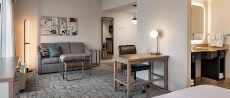 Habitación de hotel con cama, escritorio, sofá, silla, mesita, alfombra, una obra de arte en la pared, espejo y baño visible a través de una puerta abierta.