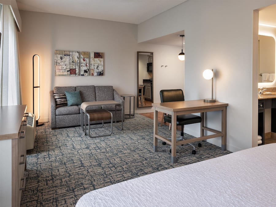 Une chambre d’hôtel comprenant un lit, un bureau, un canapé, une chaise, une petite table, un tapis, une œuvre d’art murale, un miroir et une salle de bain visible à travers une porte ouverte.