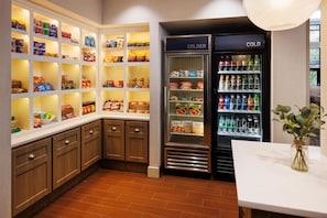 A kitchen with wooden cabinets, a built-in pantry with snacks, and a refrigerator with beverages. - Homewood Suites by Hilton St Louis Park at West End (St. Louis Park)