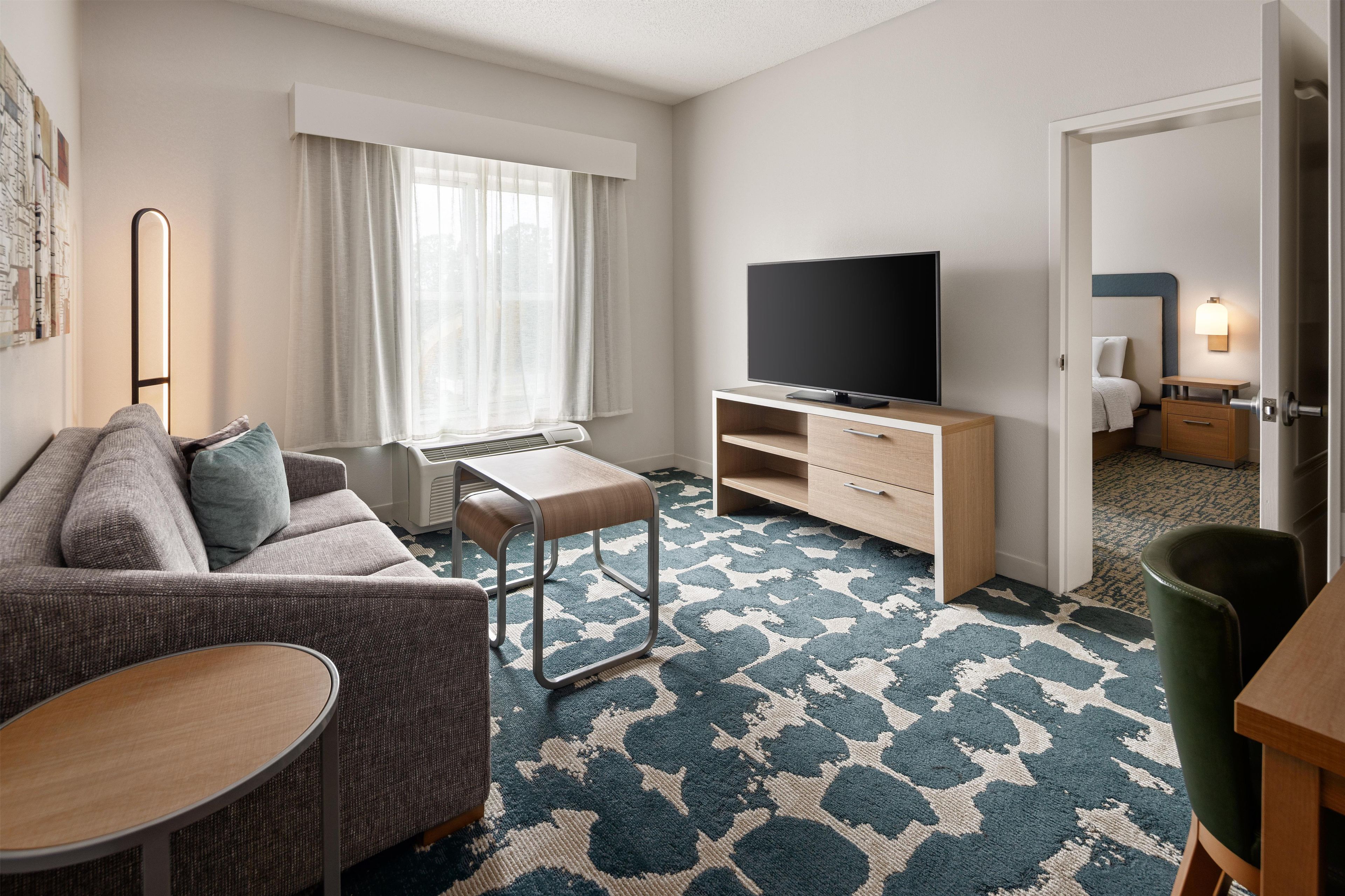 A modern hotel room with a sofa, a small table, a TV on a wooden stand, and a patterned carpet.