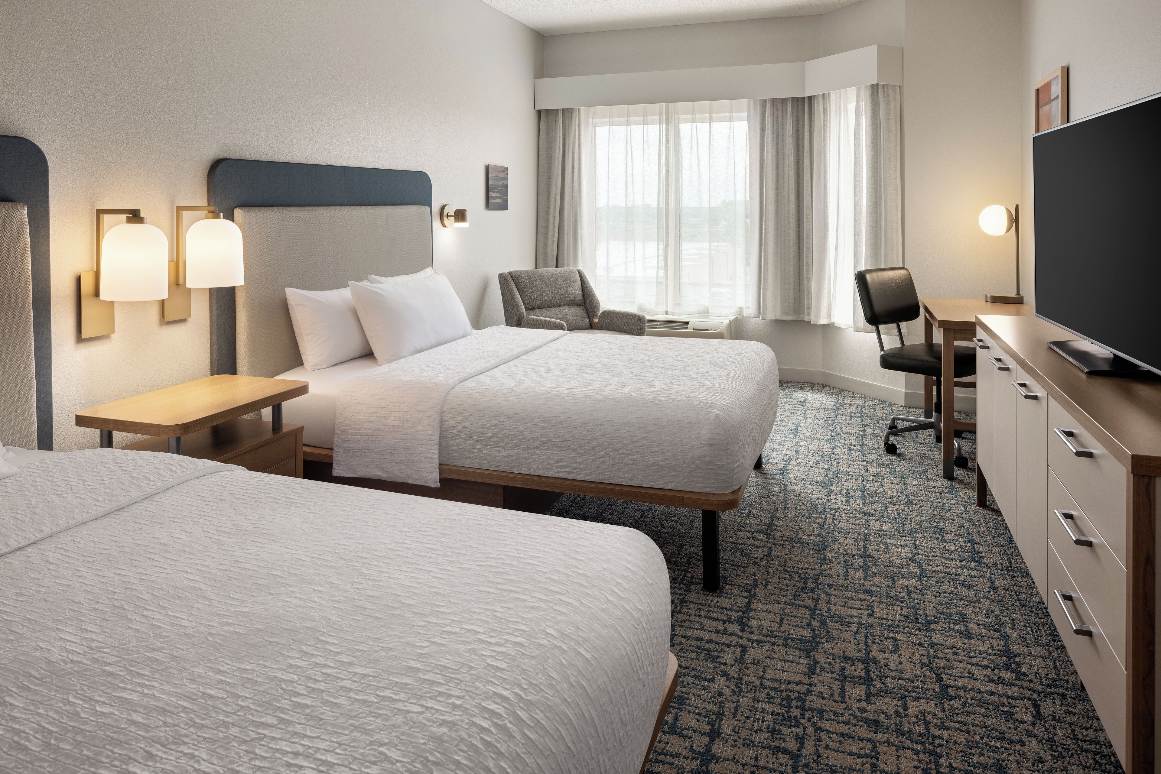 A hotel room with two beds, a desk, a chair, a television, and a window with curtains.