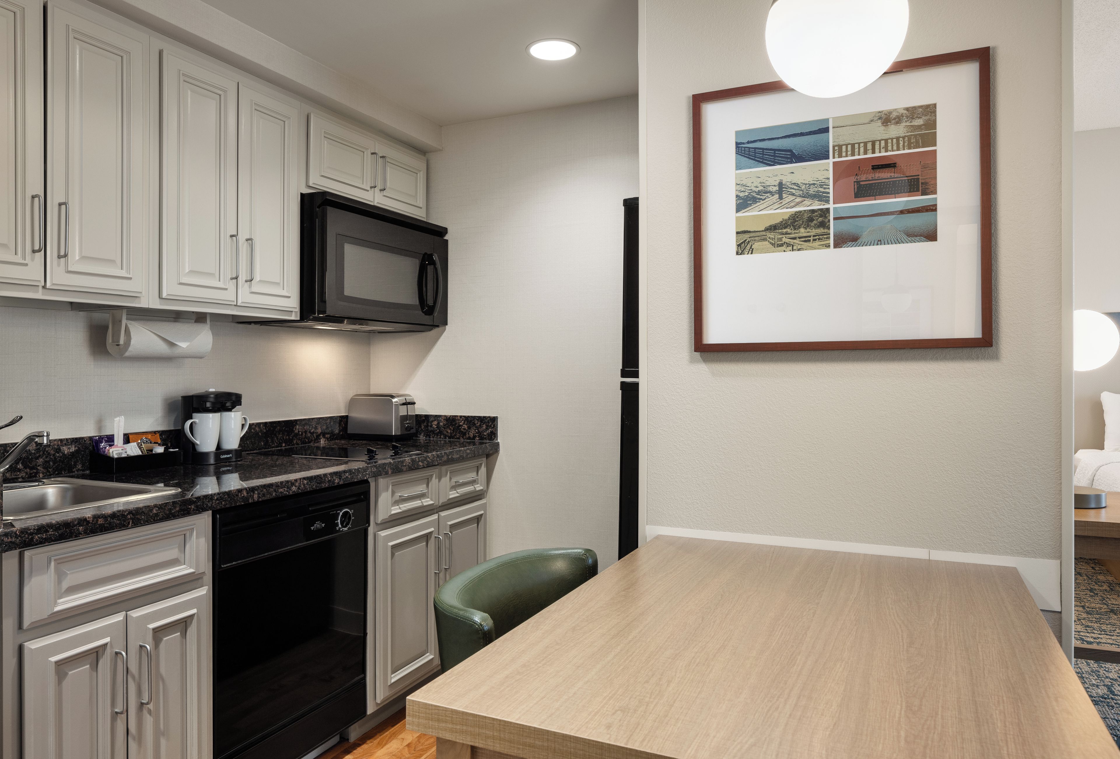 Una cocina moderna con armarios blancos, un microondas negro, una cafetera y un tostador. Hay un cuadro en la pared y una mesa de comedor con una silla.
