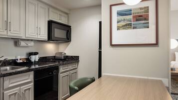 Una cocina moderna con armarios blancos, un microondas negro, una cafetera y un tostador. Hay un cuadro en la pared y una mesa de comedor con una silla.
