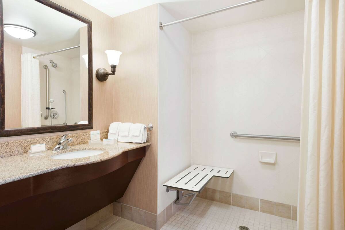 A modern bathroom with a granite countertop sink, a large mirror, a towel rack, and a shower area with a grab bar.