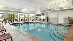 Una piscina interior con agua cristalina, rodeada de sillones de descanso y mesas, ventanas amplias y suelo de baldosas.