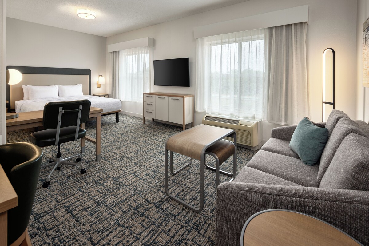 A hotel room with a bed, a desk, a chair, a sofa, a TV, and a window with curtains.