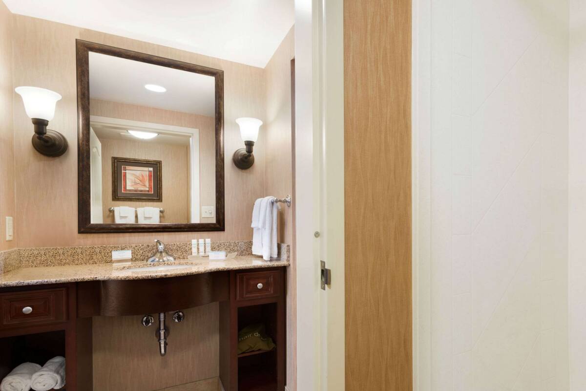 A hotel bathroom with a large mirror, two wall-mounted lights, a sink with a vanity, and a towel rack.