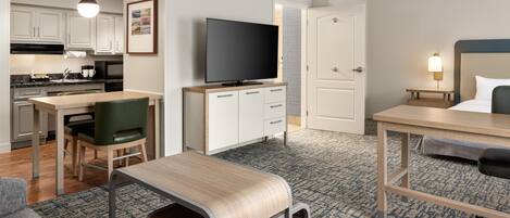 Una habitación de hotel compacta con kitchenette, televisor de pantalla plana, cama con cabecera, pequeño escritorio y zona de estar con sillón y reposapiés.