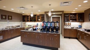 Una cocina moderna con encimeras de granito, gabinetes de madera y electrodomésticos de acero inoxidable. Hay varios dispensadores de café y una gran variedad de utensilios y electrodomésticos.