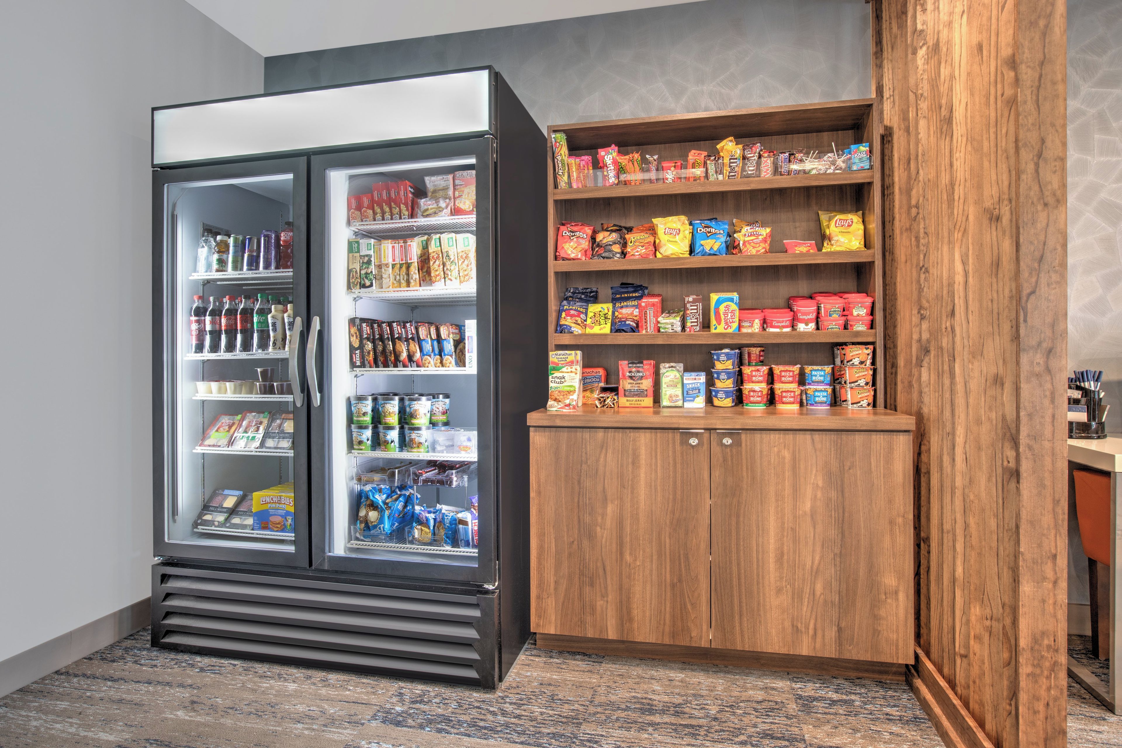 Una habitación con un refrigerador lleno de bebidas y aperitivos, y un mueble de madera con distintos alimentos.