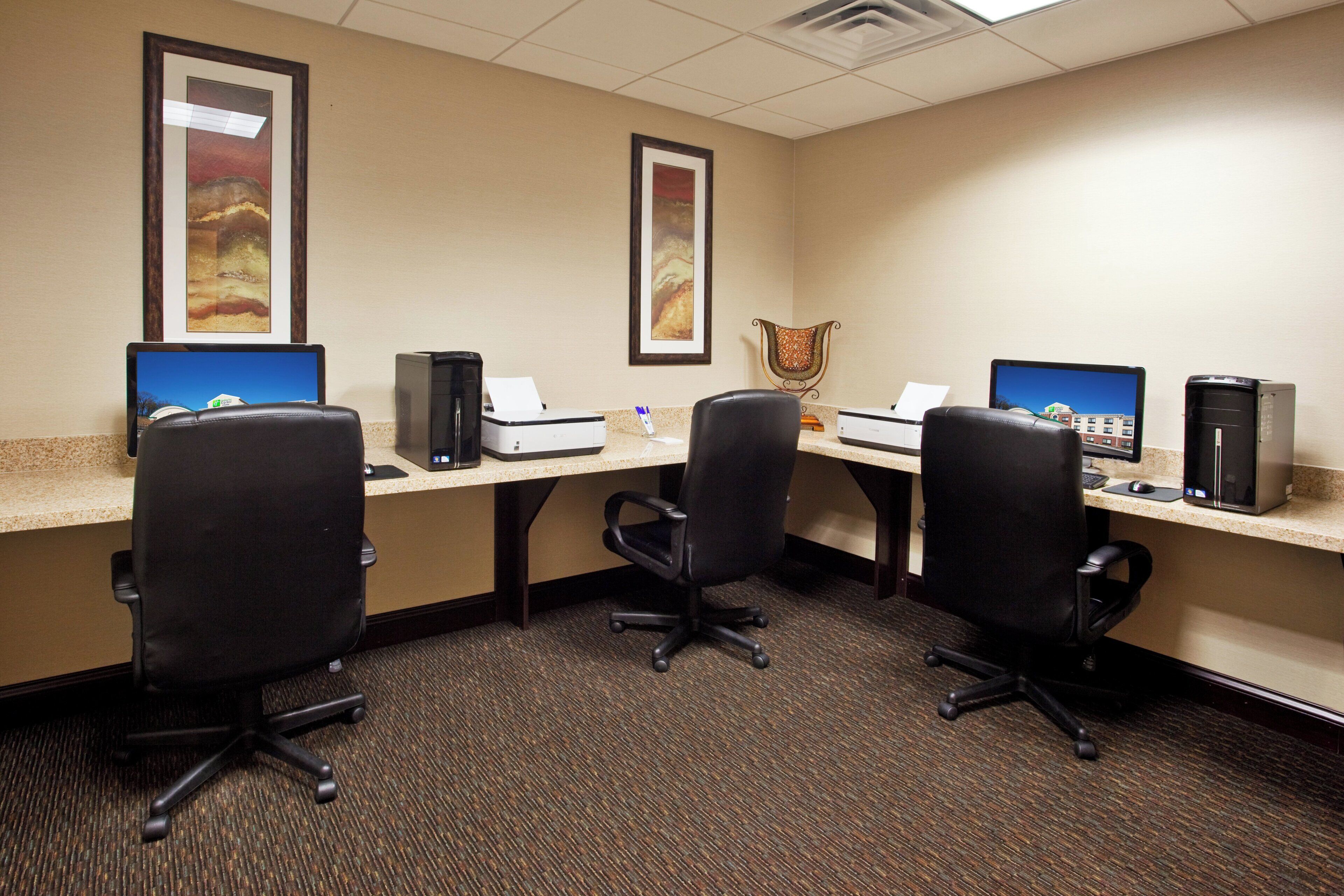 A hotel room with three desks, each with a computer and a printer, and a chair in the center.