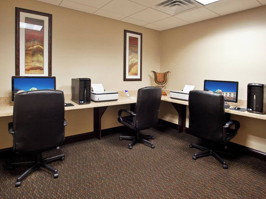 A hotel room with three desks, each with a computer and a printer, and a chair in the center.