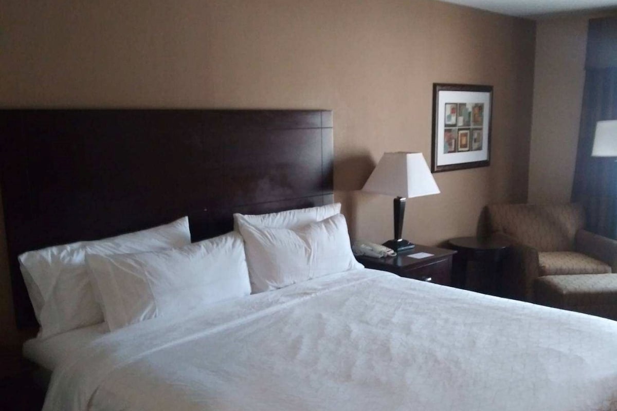 A hotel room with a large bed, a nightstand, a chair, and a framed picture on the wall.