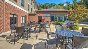 Un patio al aire libre con mesas y sillas, una parrilla y un edificio de paredes de ladrillo y ventanas grandes.