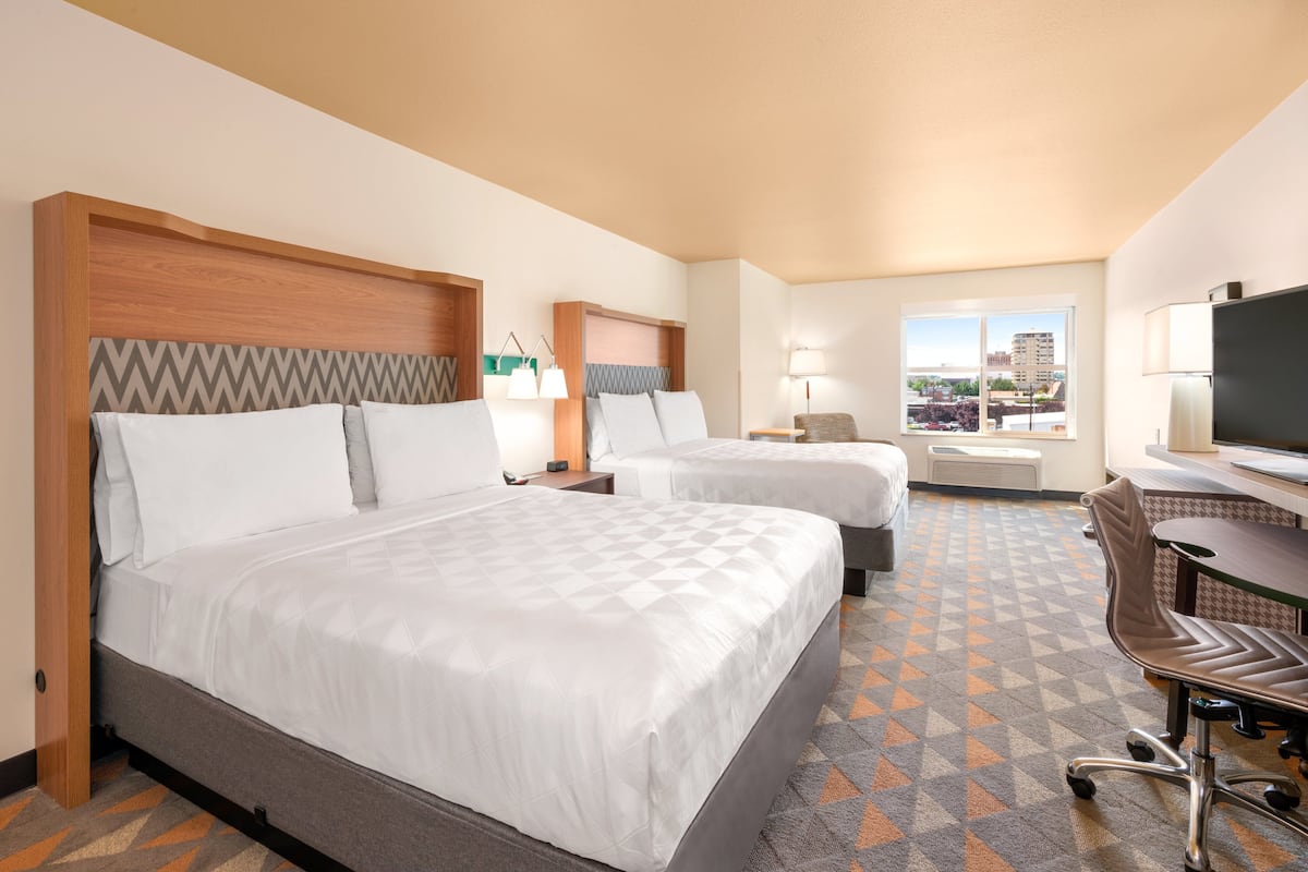 A hotel room with two beds, a desk, a chair, a TV, and a window with a city view.