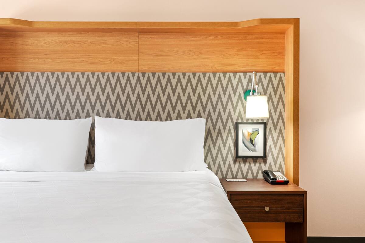 A neatly made bed with white linens, a wooden headboard, a nightstand with a lamp, and a framed picture.