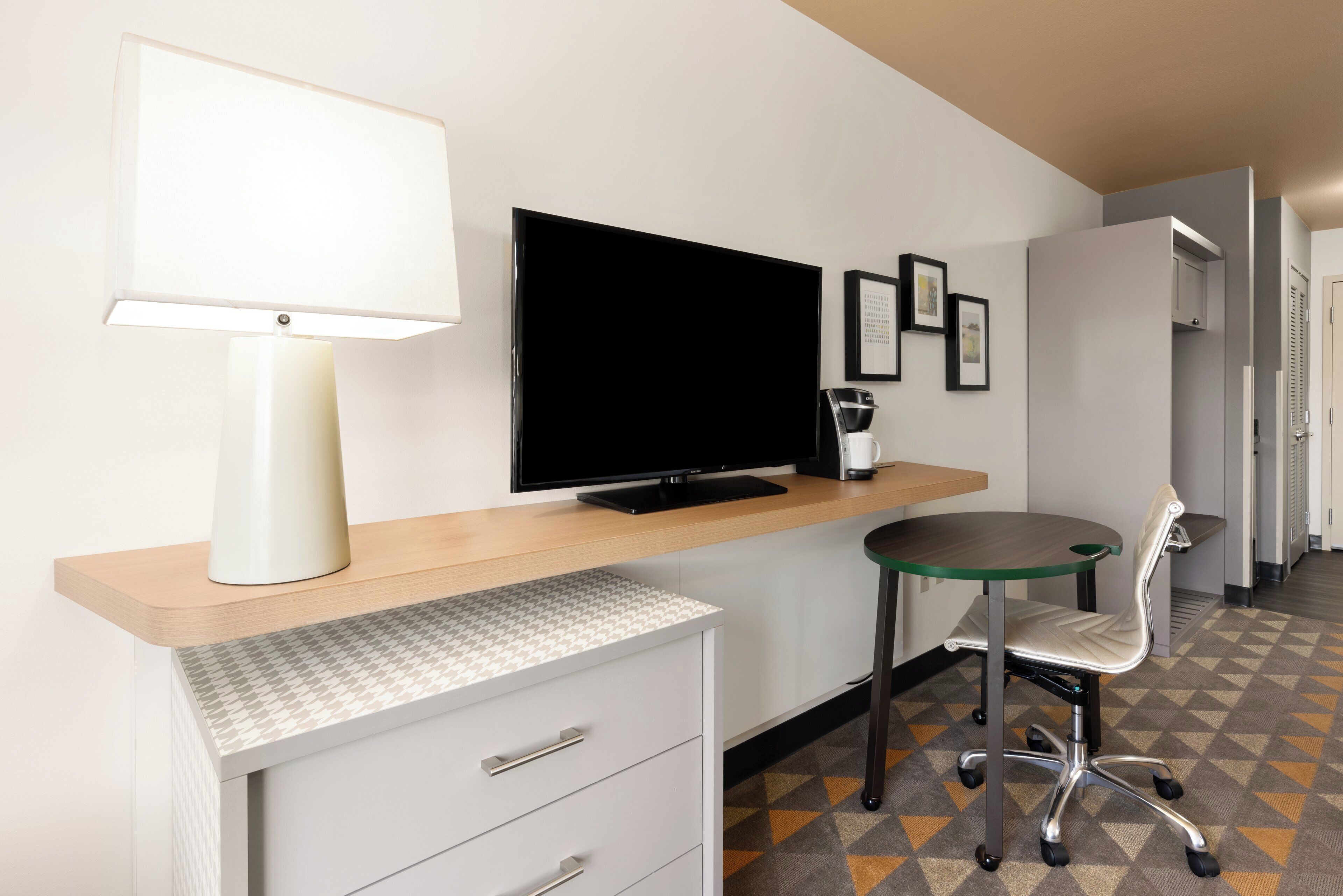 A hotel room with a desk, a flat-screen TV, a lamp, a coffee maker, a chair, and a patterned carpet.