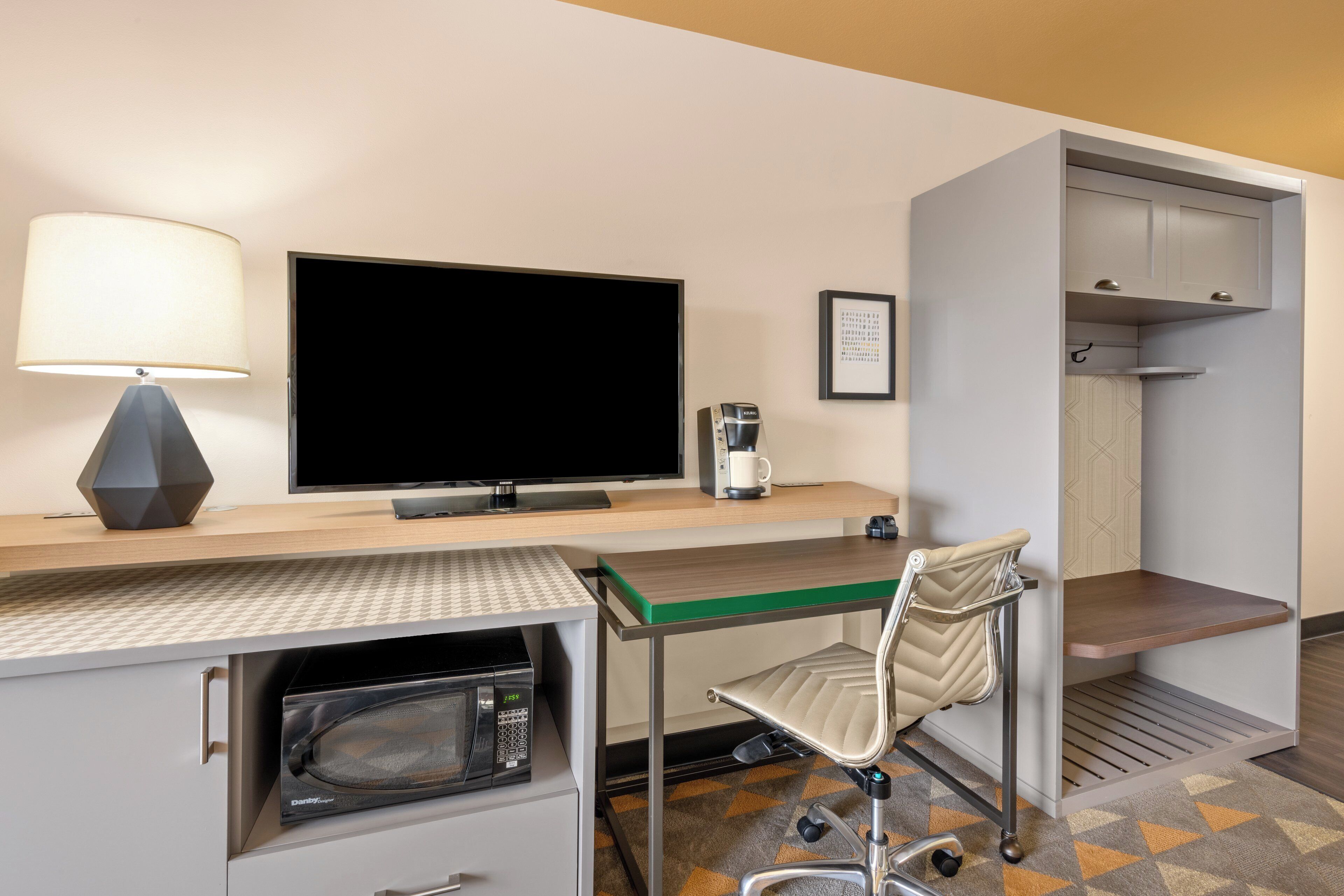 A hotel room with a flat-screen TV, a coffee maker, a microwave, a chair, and a desk.