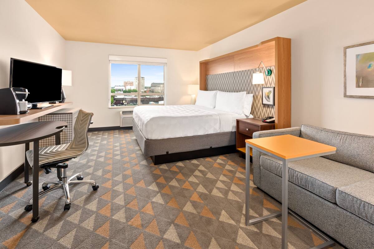 A hotel room with a bed, a desk with a computer, a sofa, and a large window with a city view.