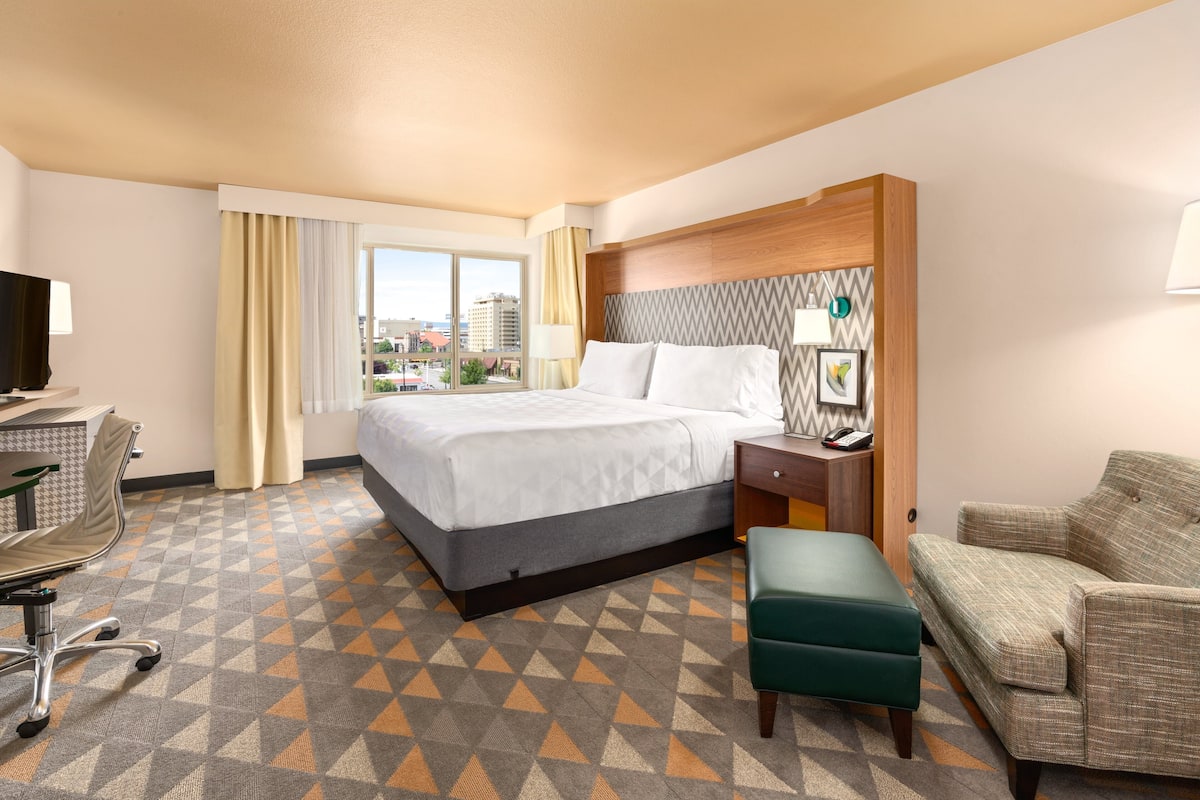 A hotel room with a large bed, a desk with a chair, a sofa, and a view of the city through the window.