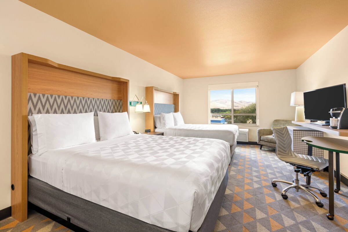 A hotel room with two beds, a desk, a chair, a TV, and a window with a view.
