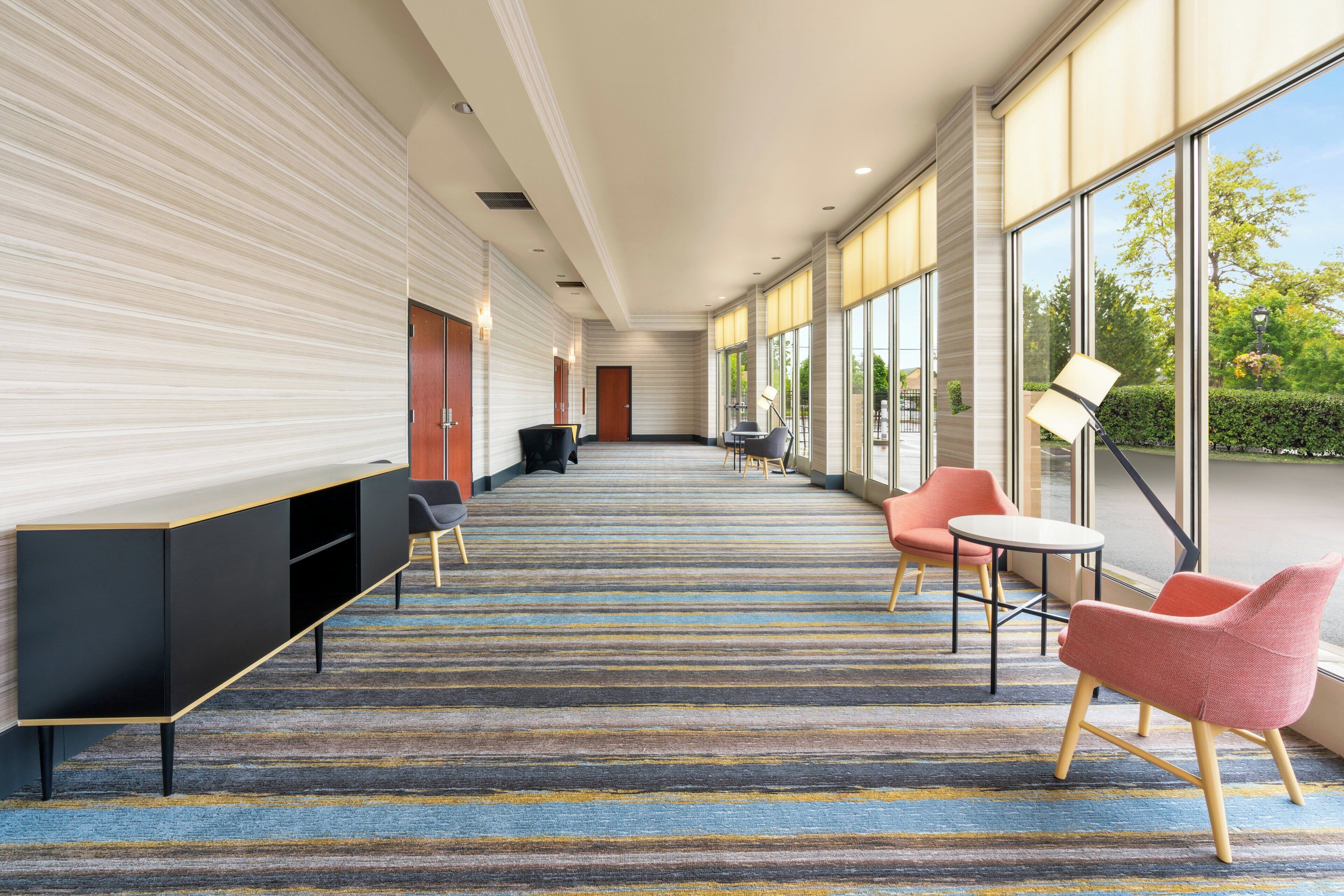 A hotel corridor with a striped carpet, seating area with chairs and tables, and large windows offering a view of greenery.