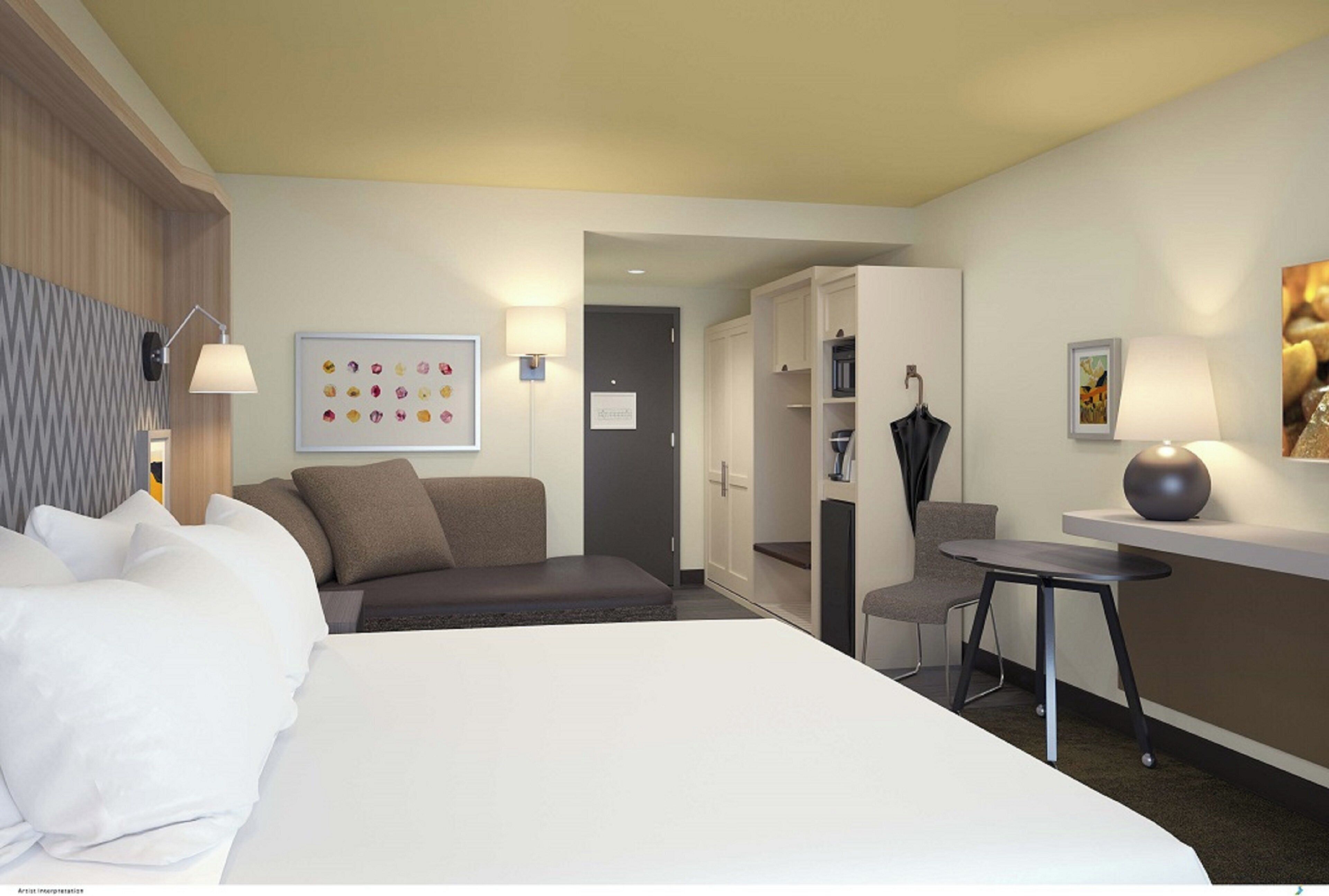 A hotel room with a bed, a desk, a chair, a wardrobe, and a wall with a colorful artwork.