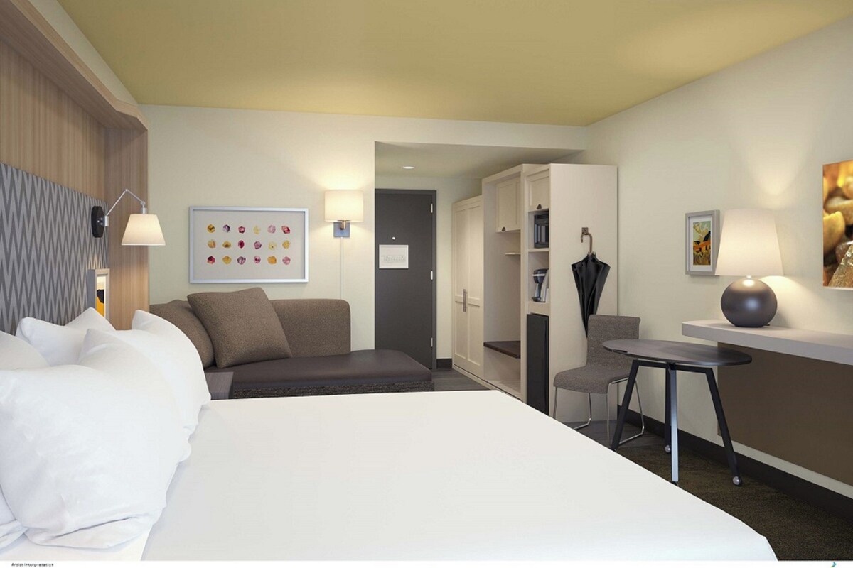 A hotel room with a bed, a desk, a chair, a wardrobe, and a wall with a colorful artwork.