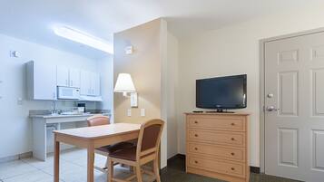 A compact room with a kitchenette, a dining table with chairs, a television on a dresser, and a door.