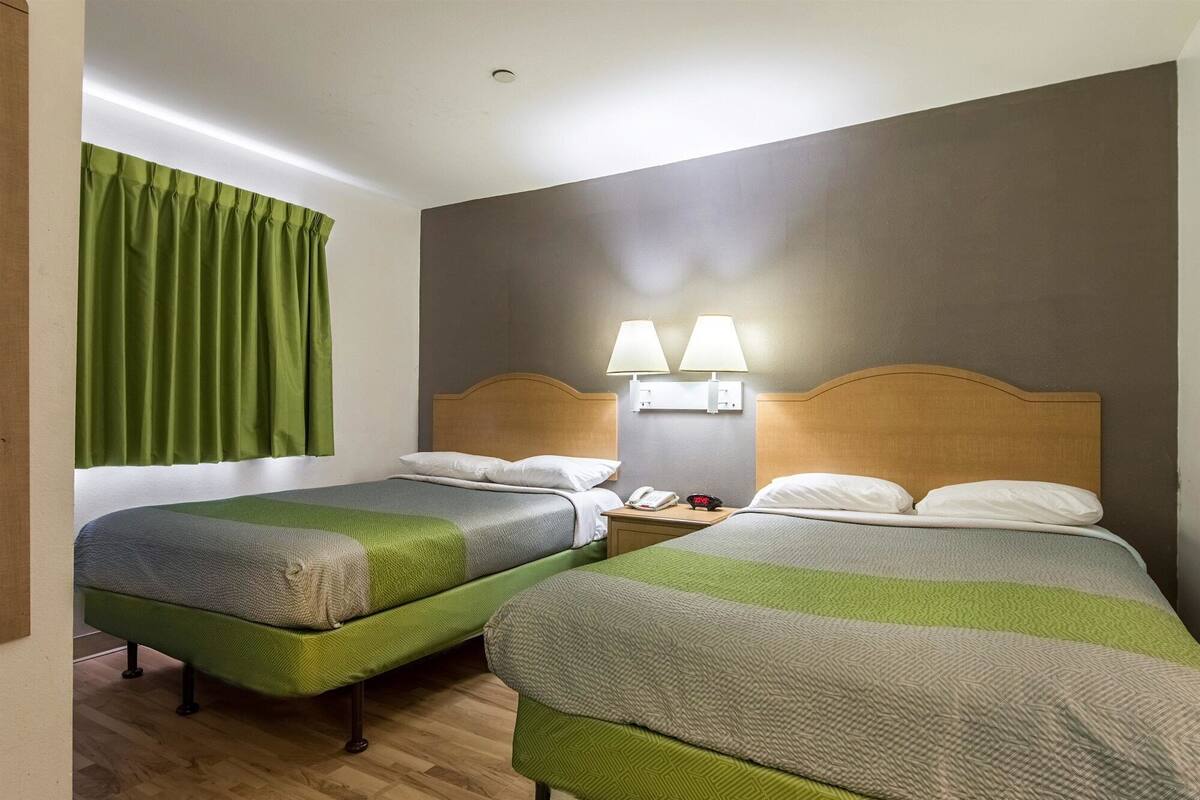 A hotel room with two beds, each with a green and grey patterned bedspread, a wooden headboard, and a nightstand with a lamp and a phone.
