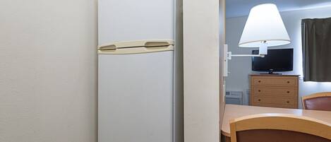 A compact room with a white refrigerator, a wooden desk with a lamp, a television, and a chair.