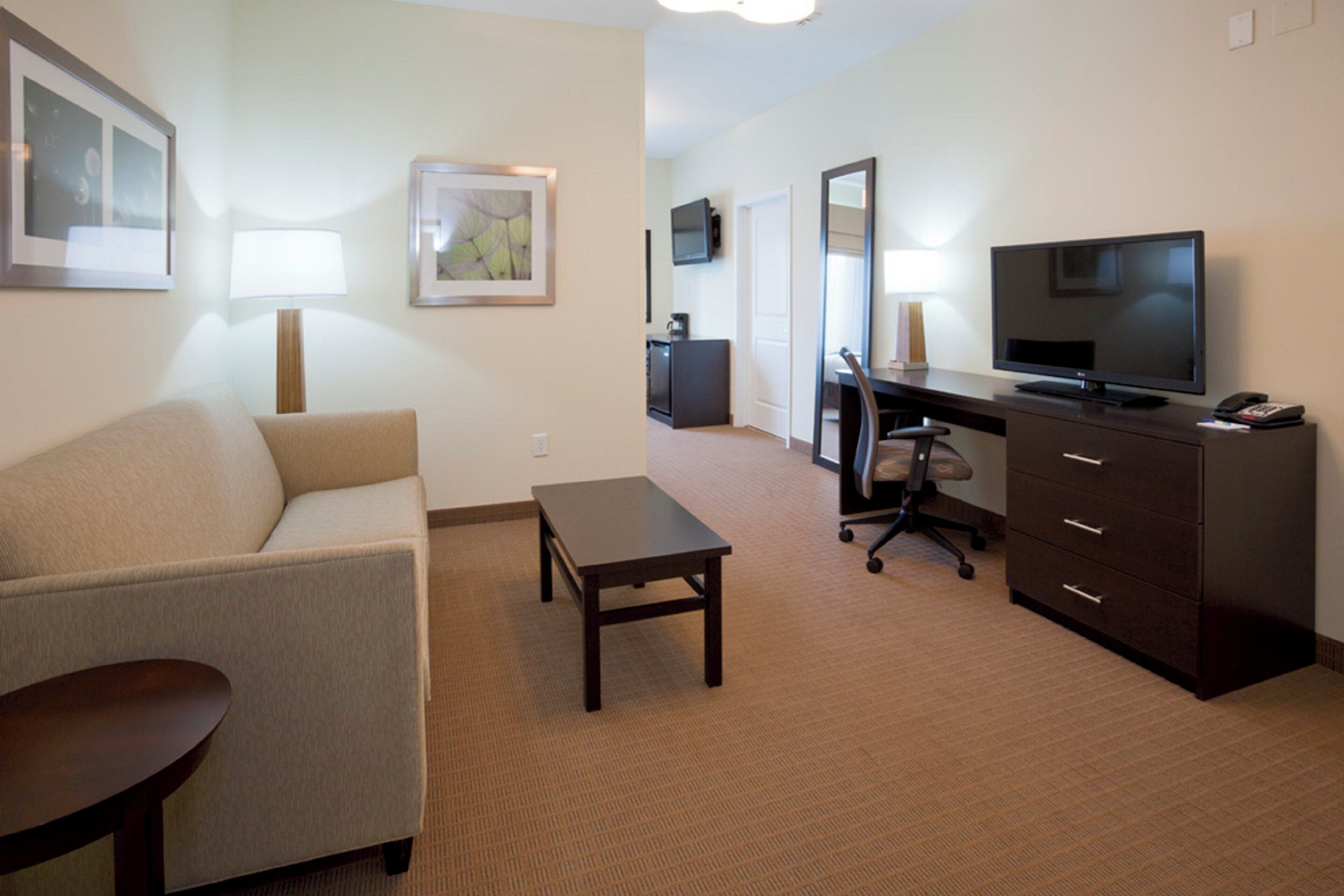 A hotel room with a sofa, a small table, a desk with a chair, a television, and a dresser.