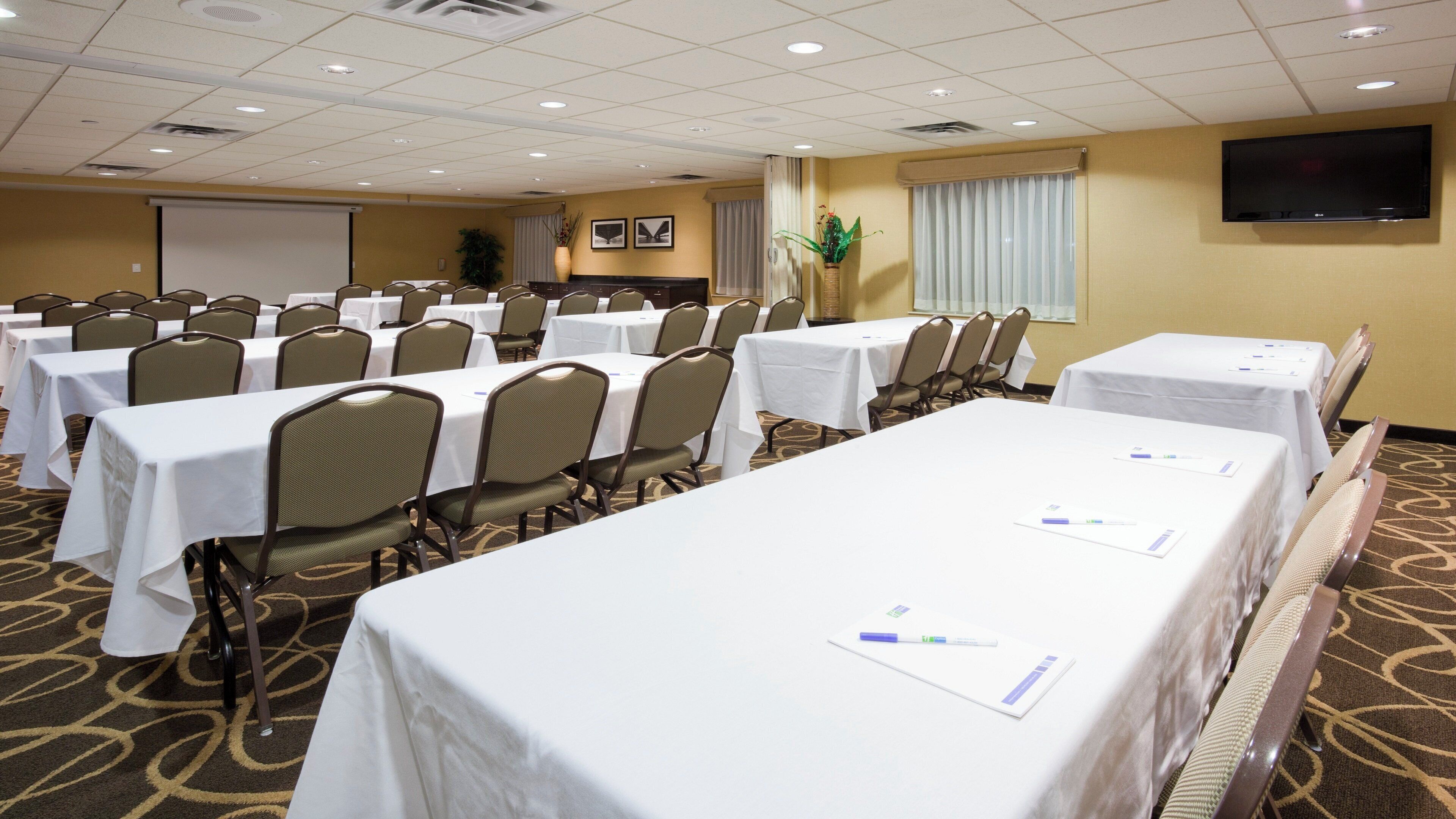 A conference room with multiple tables and chairs, a projector screen, and a television on the wall.